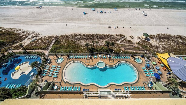 Vue aérienne d’un complexe hôtelier en bord de mer, avec une grande piscine, de nombreux transats et une plage aménagée avec des parasols et des personnes.