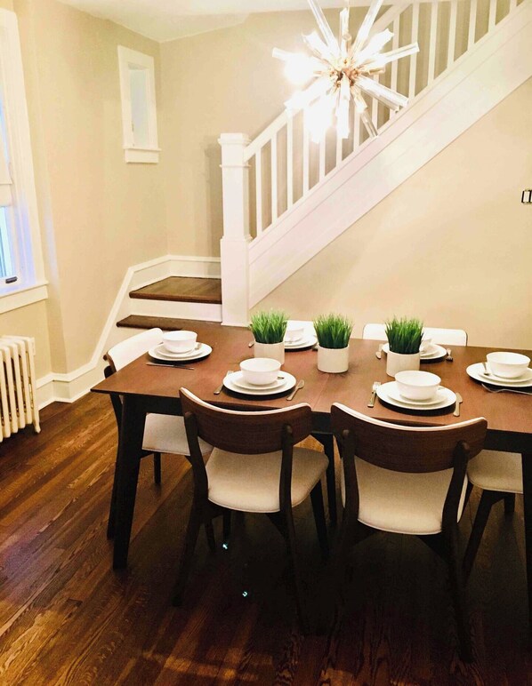 A dining room with a wooden table set for four, white chairs, and a staircase in the background. - Hilltop Haven @ Hampton Heights (Reading)