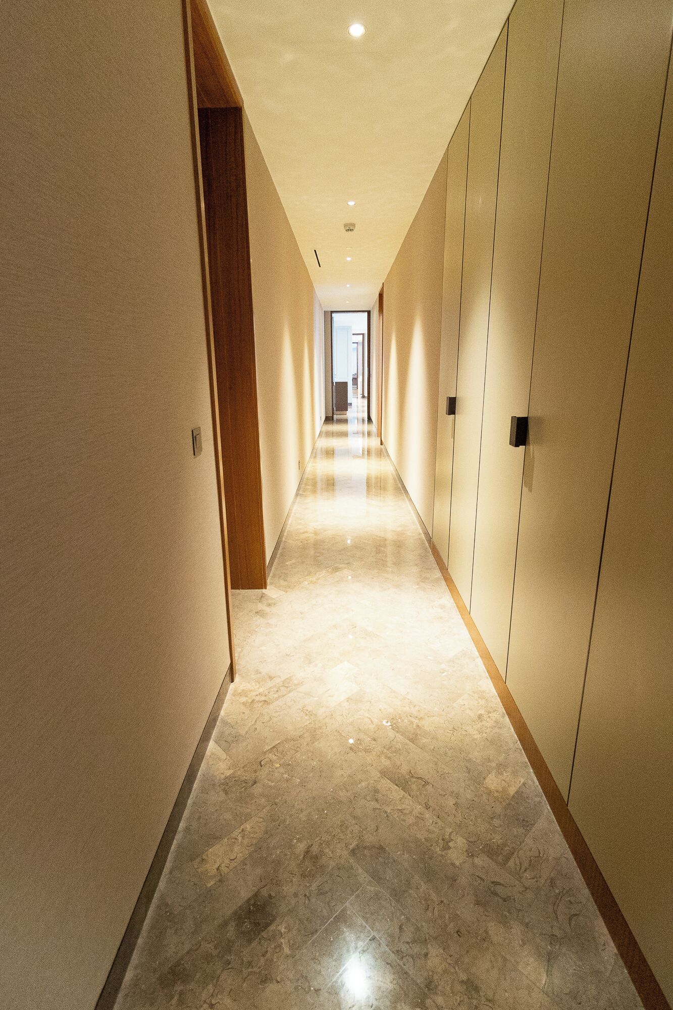A long, narrow hallway with a polished marble floor, wooden paneling on the walls, and a door at the end leading to another room.