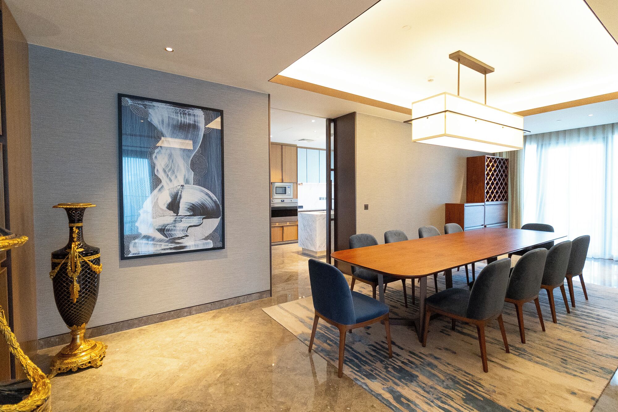 A modern hotel room with a large dining table, comfortable seating, a large painting, and a decorative vase.