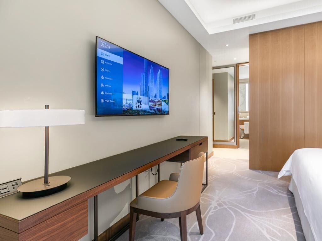 A hotel room with a bed, a desk with a chair, a flat-screen TV mounted on the wall, and a wooden wardrobe.