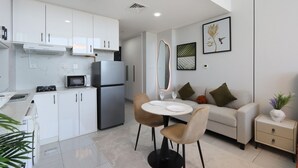 Eine moderne Küche mit weißen Schränken, einer eingebauten Mikrowelle und einem Edelstahl-Kühlschrank. Es gibt eine Essecke mit einem runden Tisch und zwei Stühlen. Außerdem sind eine Couch und ein Nachttisch mit Lampe zu sehen.