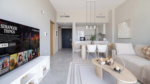 A modern living room with a flat-screen TV displaying a movie, a dining area with a table and chairs, and a sofa. - Cozy Boho Retreat One Bedroom Sanctuary (Jumeirah Village Circle)