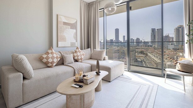 A modern living room with a large window offering a city view, a beige sofa with patterned cushions, a small coffee table, and a floor lamp. - Cozy Boho Retreat One Bedroom Sanctuary (Jumeirah Village Circle)