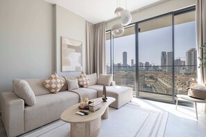A modern living room with a large window offering a city view, a beige sofa with patterned cushions, a small coffee table, and a floor lamp. - Cozy Boho Retreat One Bedroom Sanctuary (Jumeirah Village Circle)
