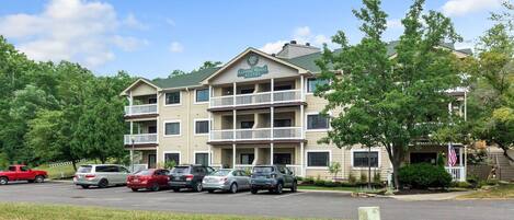 A multi-story hotel with a balcony on the second floor, a parking lot with several cars, and a flag on the building.