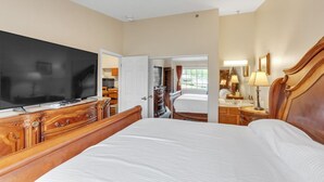 A hotel room with a large bed, a TV, a wooden dresser, and a bathroom with a mirror and lamp. - 3rd Floor Front Side Corner Penthouse Suite - 304 by Grand Wood Suites (Nashville)