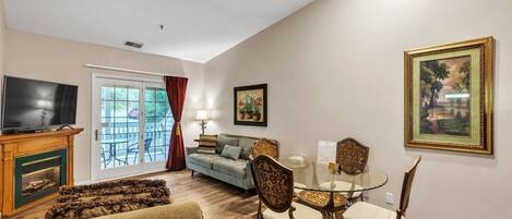 A living room with a television, sofa, and a glass table with chairs. There is a fireplace, a door leading to a balcony, and a painting on the wall.