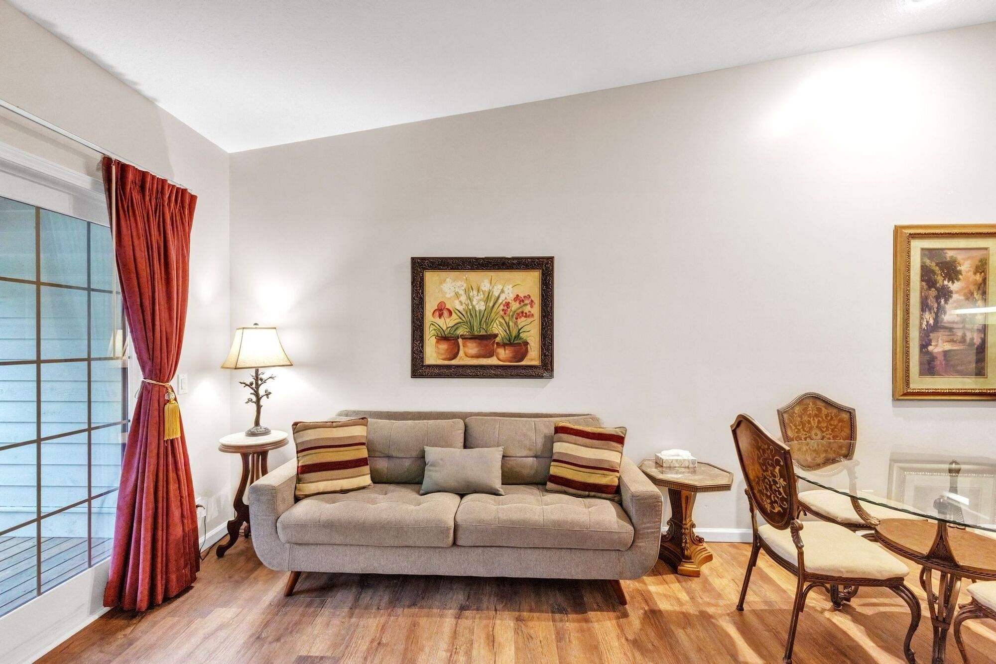 A living room with a sofa, a side table, a lamp, a framed painting, and a dining area with a chair and table.