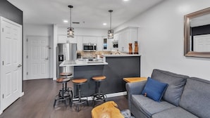 Una cocina moderna con gabinetes blancos, electrodomésticos de acero inoxidable y una isla central. Hay dos taburetes de bar y un sofá gris con un cojín azul.