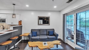 Cocina moderna con armarios blancos, una isla de madera y una zona de bar. En la sala de estar, hay un sofá gris con cojines azules, y ventanas amplias que ofrecen vistas a árboles.