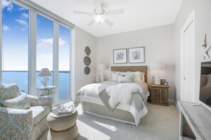 Une chambre avec une grande fenêtre, un lit, une chaise, une table de chevet et un meuble pour la télévision.