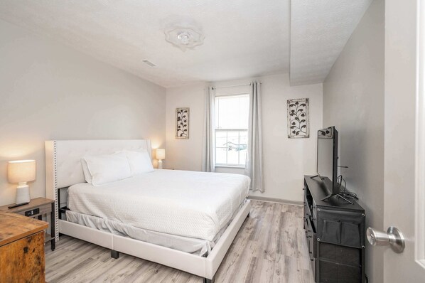 A bedroom with a white bed, bedside lamps, a wooden dresser, a mirror, and a window with curtains.