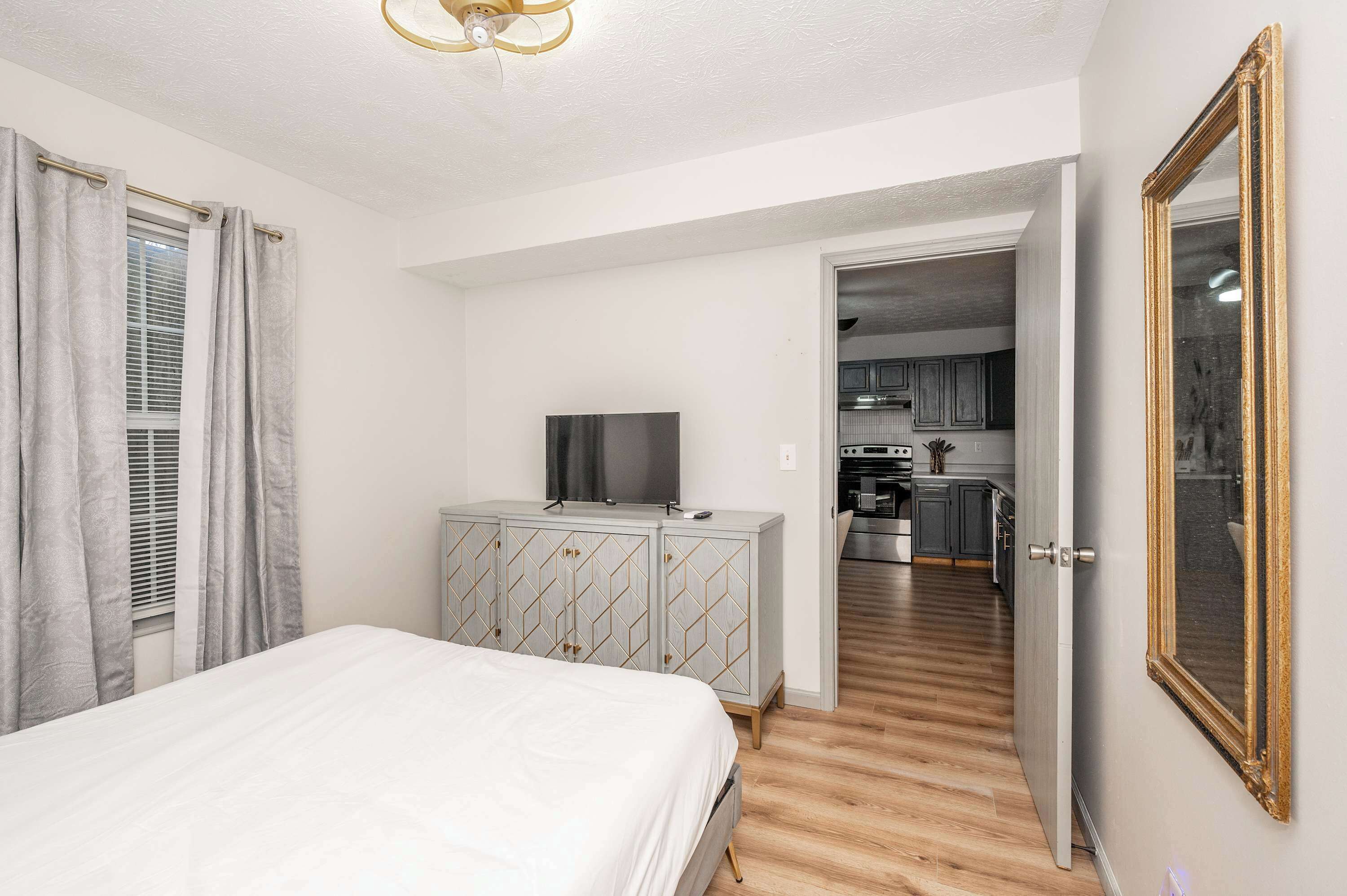 A bedroom with a bed, a TV on a cabinet, and a view into the kitchen.