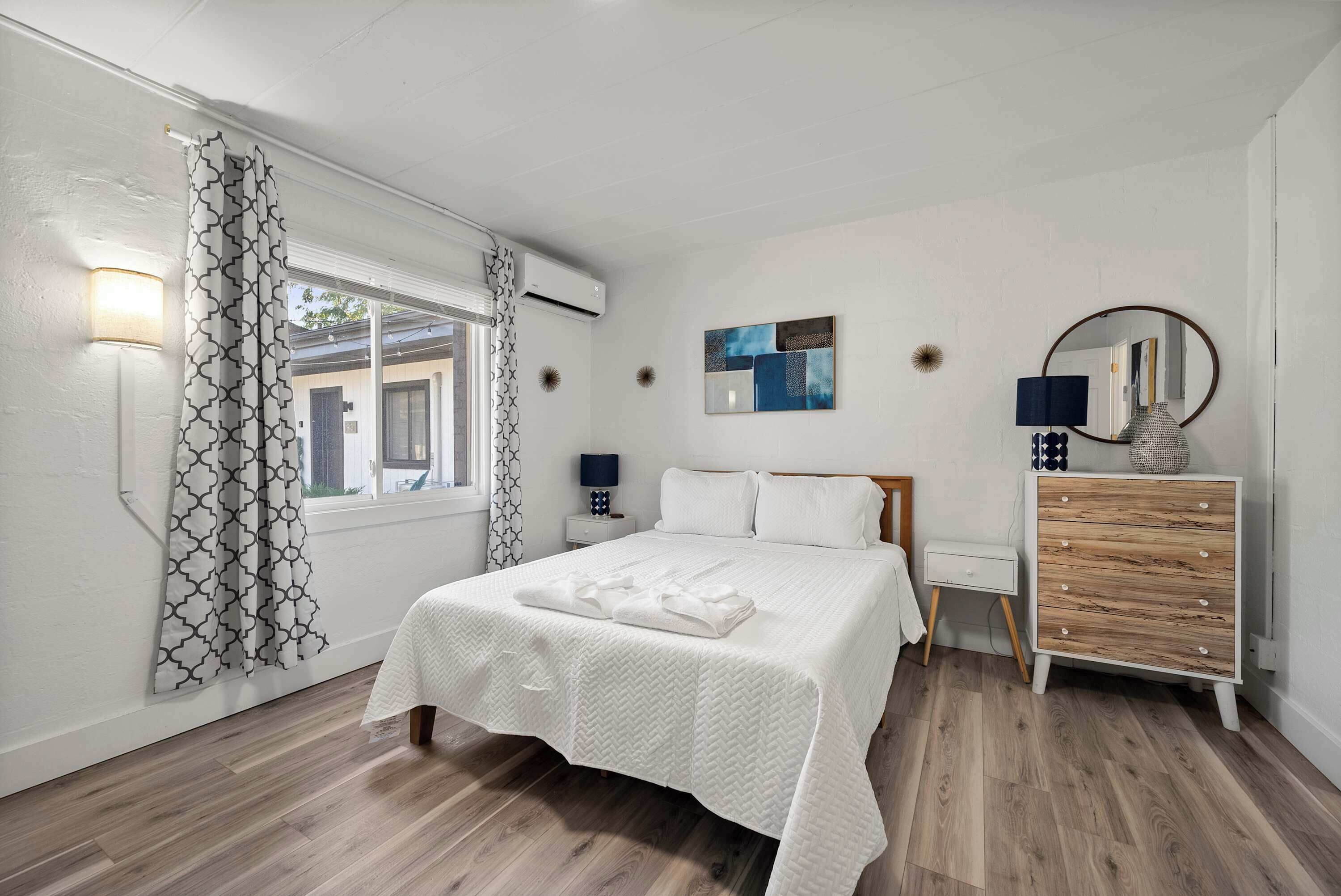 A bedroom with a bed, a dresser, a nightstand, a lamp, a mirror, and a window with curtains.