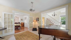 A spacious living room with a grand piano, a dining area, and a staircase leading to another level. - Classic Charm on Ashland – For 8 (Columbus)