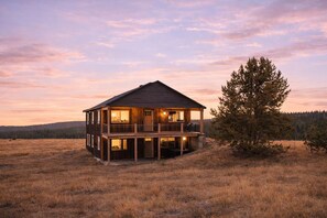 Une cabane en bois à deux étages, dotée d’un balcon, entourée d’un champ herbeux et d’une zone boisée.