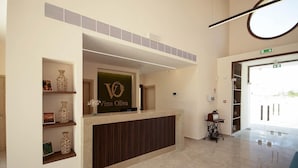 Una recepción con un letrero que dice 'Vina Olivia', un estante en la pared con objetos decorativos, una máquina de coser y un vidrio transparente que da a una puerta exterior.