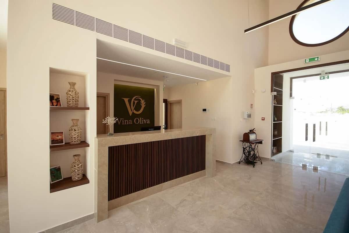 A reception desk with a sign reading 'Vina Olivia,' a wall-mounted shelf with decorative items, a sewing machine, and a clear glass door leading outside.