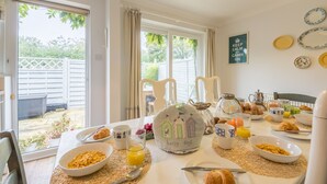 Une table de petit-déjeuner dressée avec des assiettes, des bols et des tasses, une machine à café, et une vue sur le jardin à travers de grandes fenêtres.