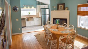 A dining area with a wooden table and chairs, a fireplace, and a kitchen in the background. - Quintessential Cape Cod condo, .4 miles to the beach (Dennis Port)