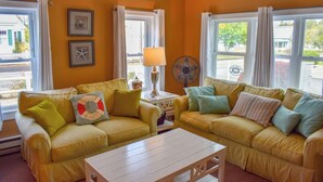 A room with two yellow sofas, a small white table, a lamp, a fan, and two windows with curtains. - Quintessential Cape Cod condo, .4 miles to the beach (Dennis Port)