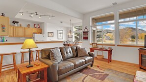 A spacious living area with a leather sofa, wooden furniture, a dining table, and large windows offering a view of the outdoors.