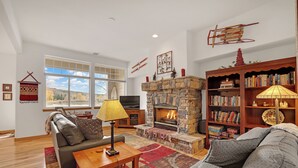 A living room with a stone fireplace, a sofa, a coffee table, a bookshelf, and a large window with a view of the outdoors.