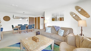 A living room with a sofa, coffee table, and dining area with chairs. There is a clock on the wall and a lamp on the side.