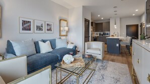 A modern living room with a blue sofa, a glass coffee table, and a kitchen in the background. - Chic Midtown Penthouse Escape - Pool, Gym, Patio with City Views (Nashville)