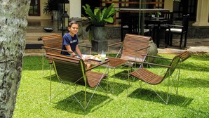 A person sitting on a wooden bench with a table and chairs in a well-maintained outdoor area with grass, a tree, and a building in the background.