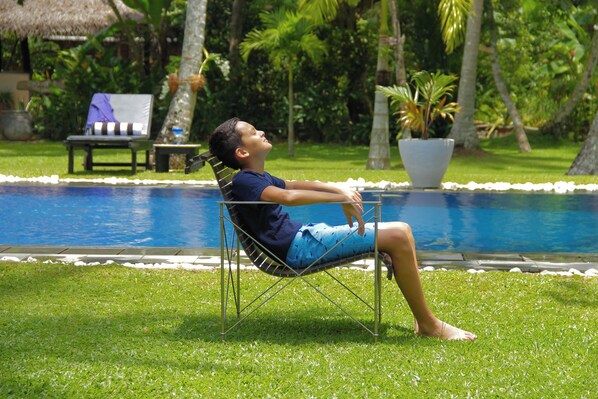 A person relaxing in a chair by a pool with a thatched roof structure and potted plants in the background.