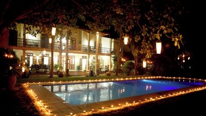 A pool surrounded by a stone deck with candles, a two-story building with balconies, and trees in the background.