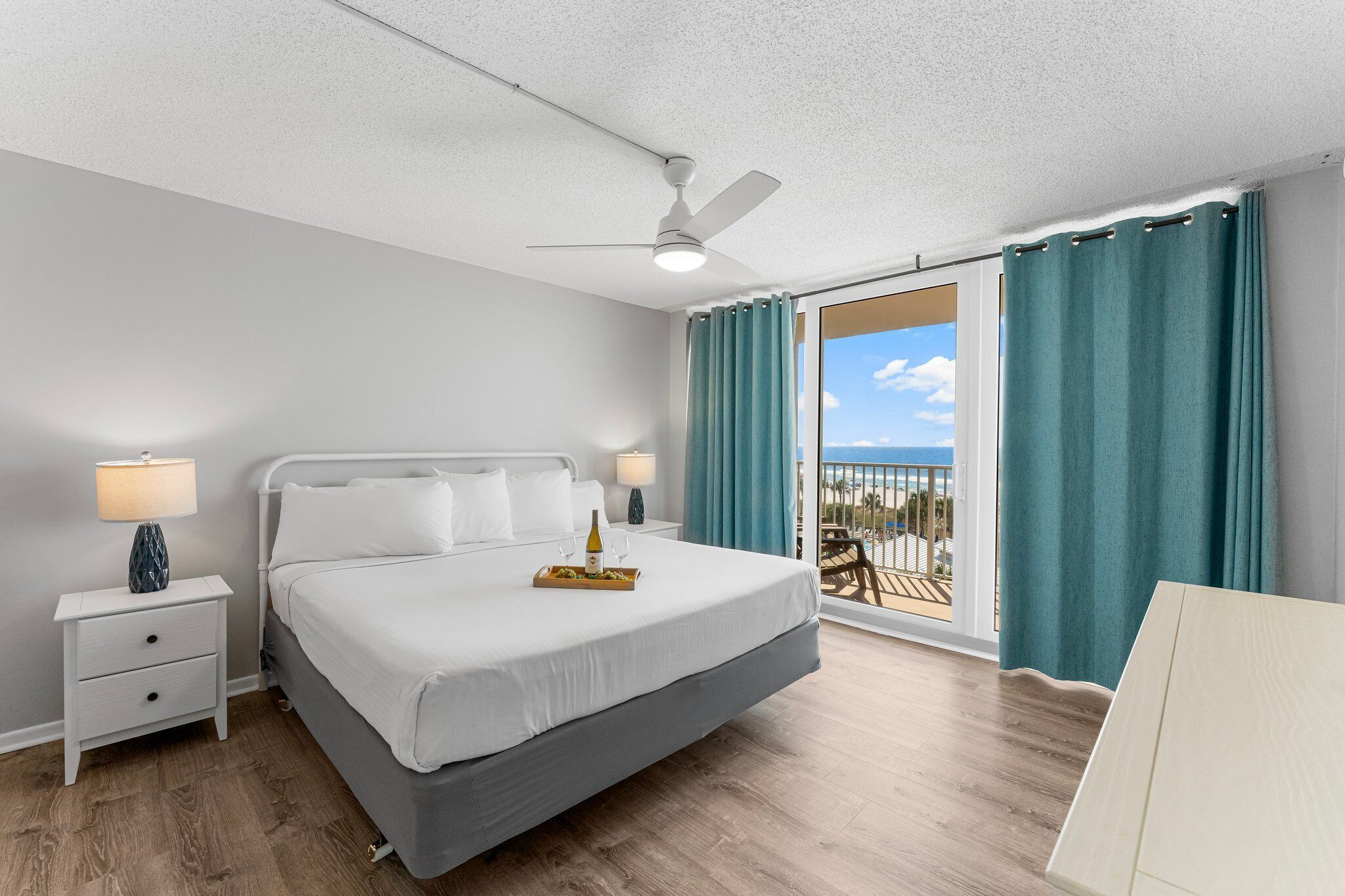 Habitación de hotel con cama, mesitas de noche, ventilador de techo, balcón con cortinas y vista al mar.