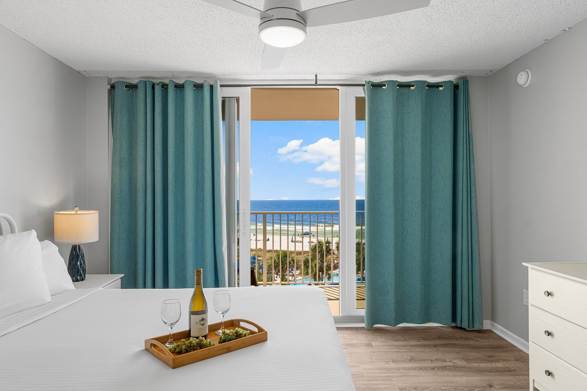 Une chambre d’hôtel avec un lit, une table de chevet avec une lampe, un plateau avec une bouteille et des raisins, et une vue sur la plage et la piscine.