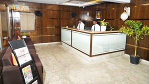 A reception area with a wooden reception desk, two staff members, a clock, and a potted plant. - Sri Brundavan Grand Inn (Anakapalle)