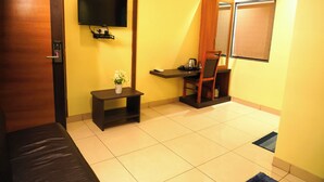 A room with a bed, a small table with a vase of flowers, a desk with a chair, a television mounted on the wall, and a window with a blind. - Sri Brundavan Grand Inn (Anakapalle)