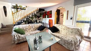 Ein Wohnzimmer mit zwei gemusterten Sofas, einem Glastisch, einer Treppe und Blick ins Freie.