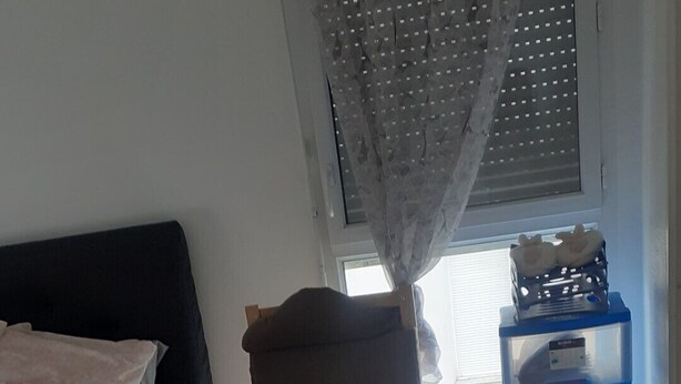 Une chambre à coucher avec un lit, une chaise, un panier en osier et une fenêtre avec des stores.