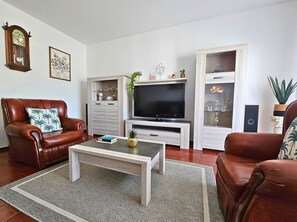 Un salon avec un canapé en cuir marron, une table basse, un téléviseur posé sur un meuble en bois, une bibliothèque et une horloge au mur.