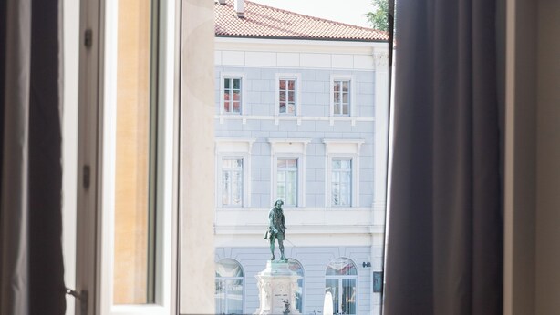 A room with a window overlooking a building with a statue and a fountain.
