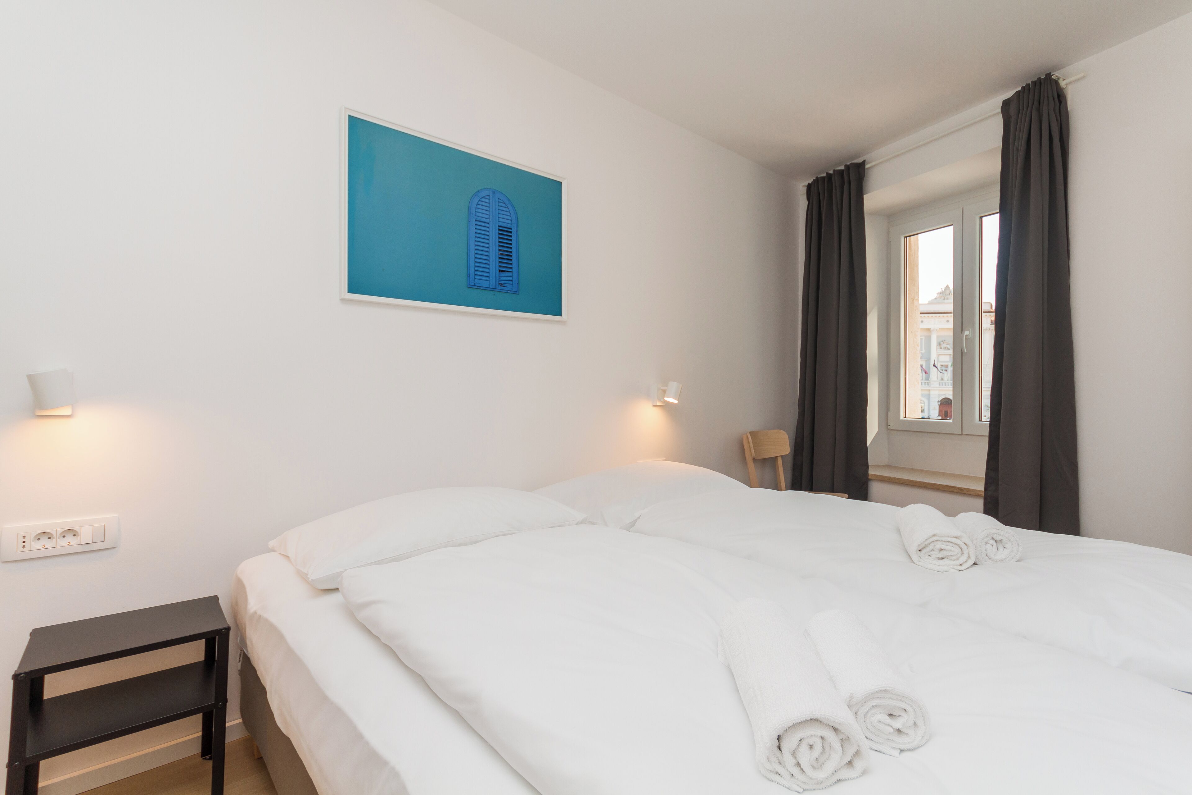 Una cama bien hecha con sábanas blancas y toallas enrolladas, una mesita de noche pequeña, una lámpara de pared y una ventana con cortinas.