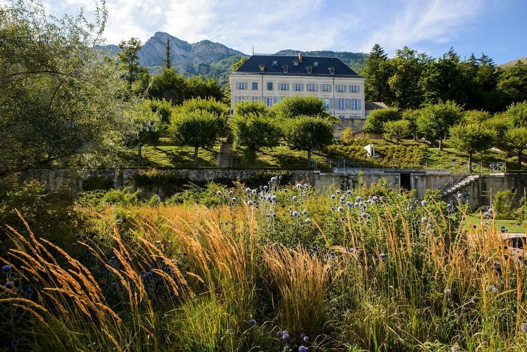A large building with a symmetrical facade, surrounded by lush greenery and a stone bridge.