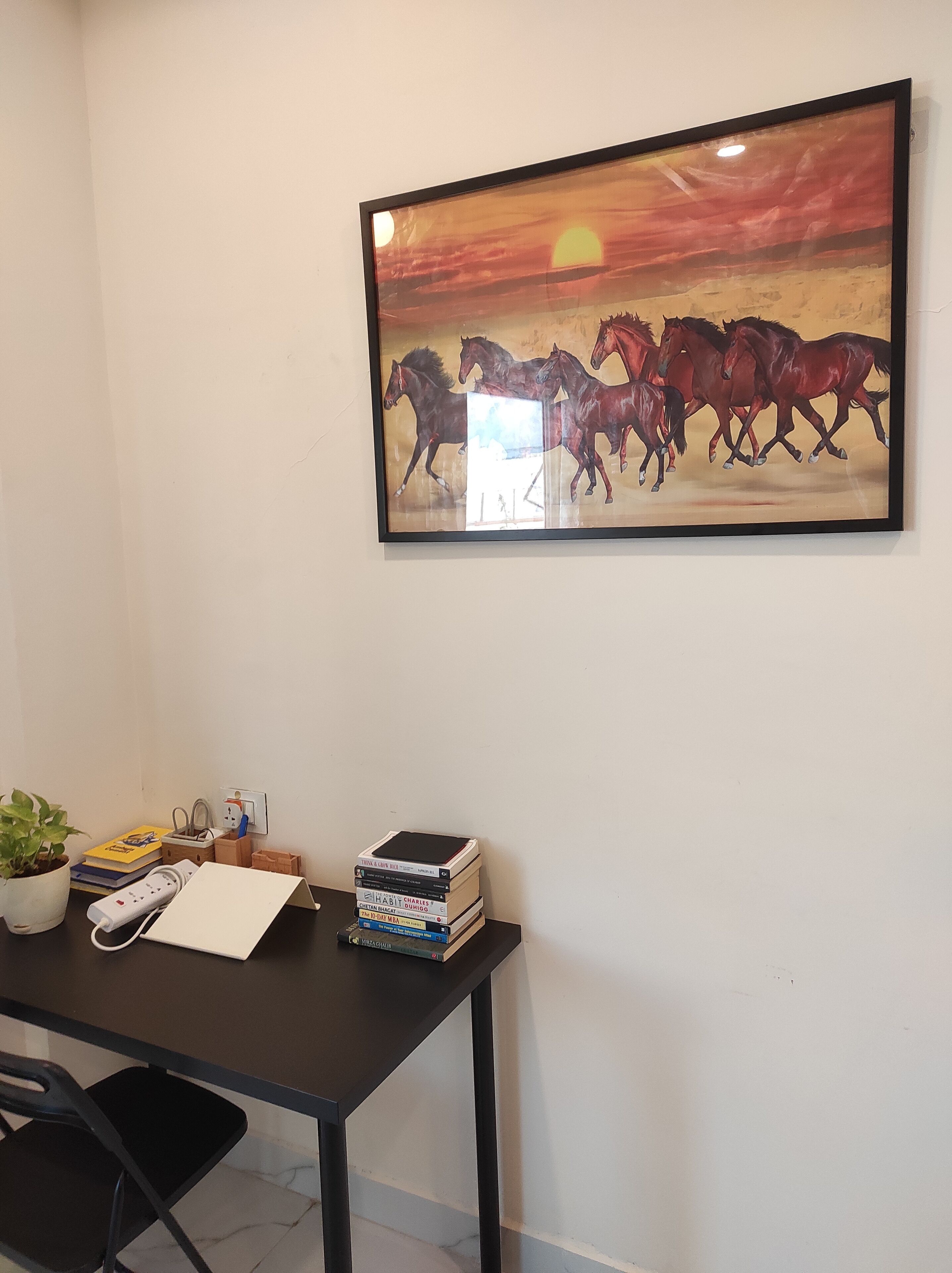 A room with a framed painting of horses, a desk with books, a power strip, and a potted plant.