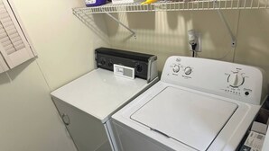 Un lavadero con dos lavadoras y una secadora, un estante con productos de limpieza y un cesto en el suelo.