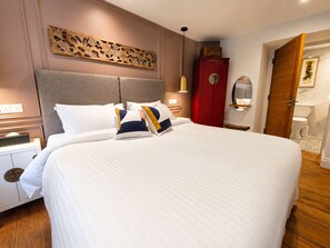 Una cama bien hecha con ropa de cama blanca y almohadones decorativos, un cabecero de madera, un armario rojo y una obra de arte colgada en la pared.