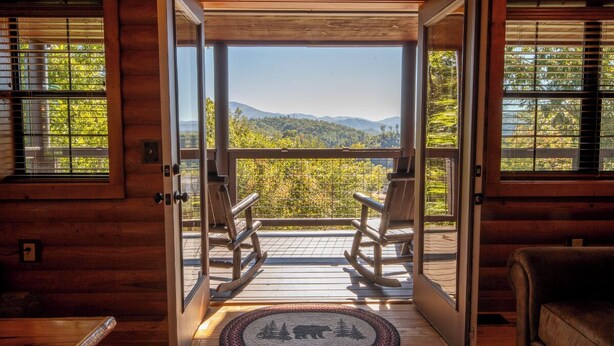 A wooden cabin with a balcony, rocking chairs, and a view of mountains and trees.