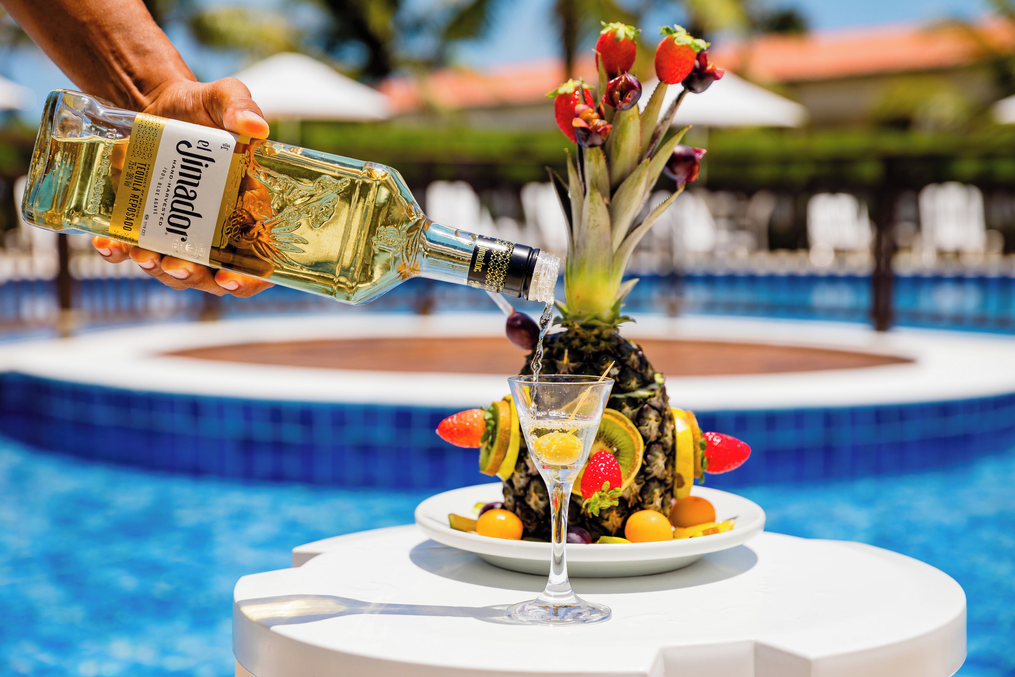 A person pouring a drink from a bottle onto a glass. There is a plate with fruits and a pineapple on a table near a pool.