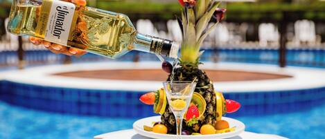 A person pouring a drink from a bottle onto a glass. There is a plate with fruits and a pineapple on a table near a pool.