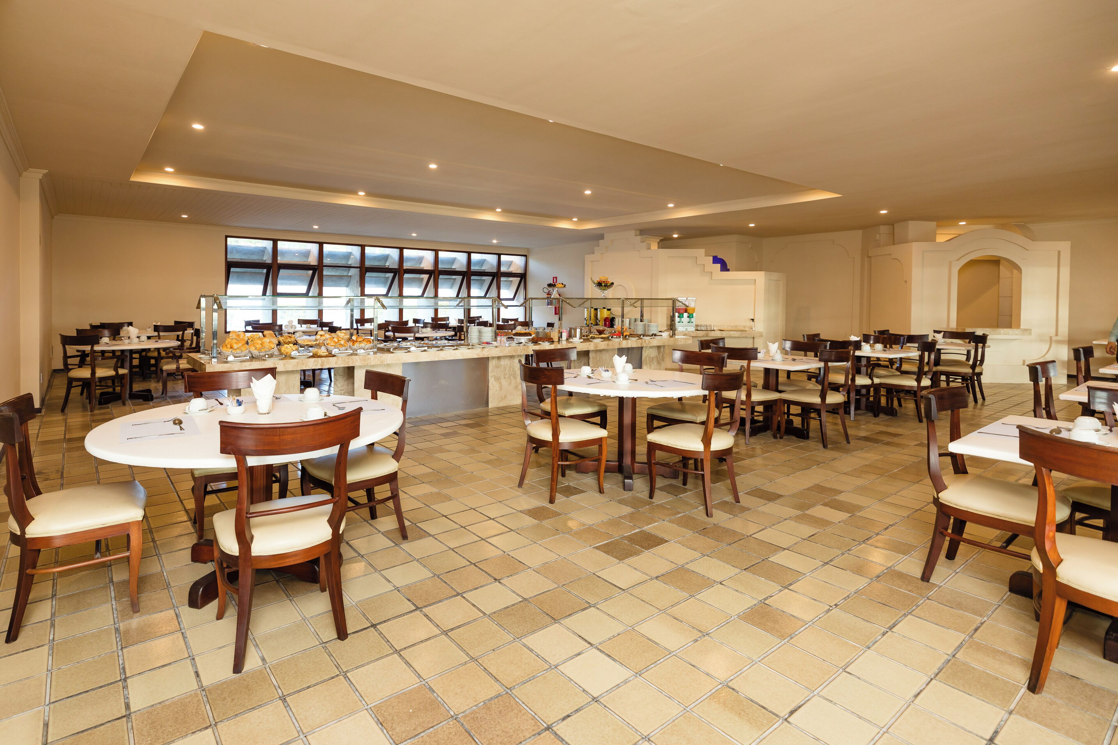 A spacious dining area with multiple round tables and chairs, a central buffet, and large windows providing natural light.